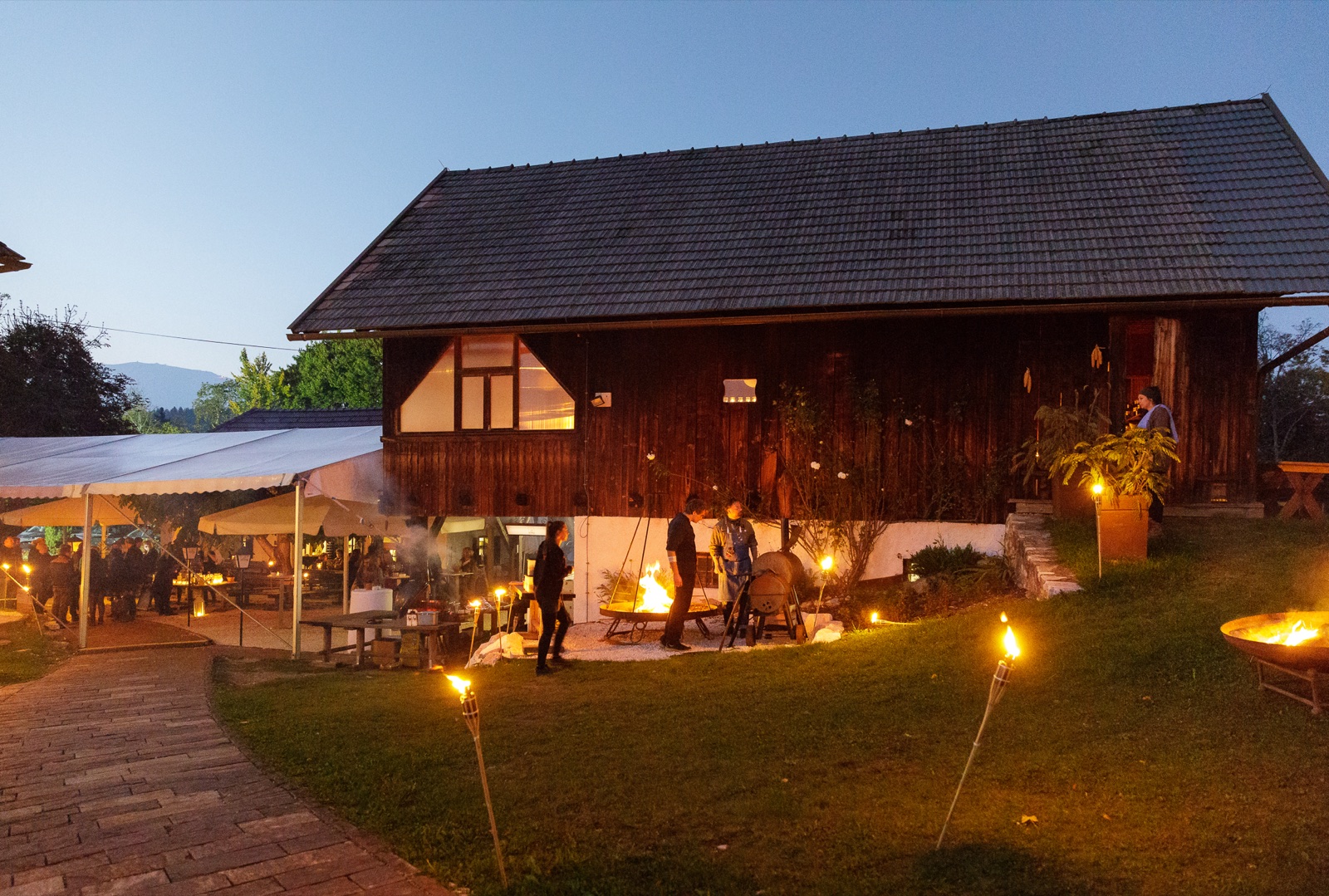 Eine Eventlocation am Abend mit Feuerstellen, Außenküche und beleuchtetem Garten.