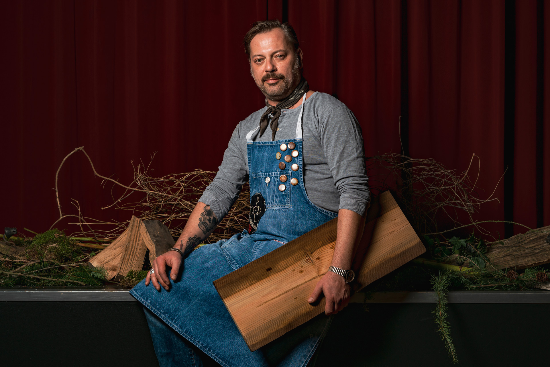 Kai Neumann sitzt vor einem dunkelroten Vorhang zwischen Holz und Zweigen.
